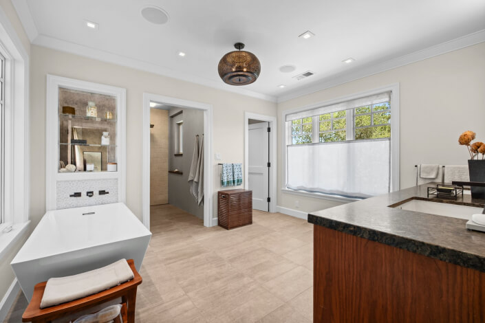 Spacious modern bathroom featuring a white bathtub, walk-in shower, large window, wooden accents, and granite countertop. The neutral color palette and stylish decor contribute to a calm and inviting atmosphere.