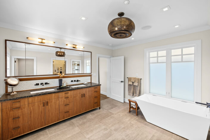 Modern bathroom featuring a wooden vanity, expansive mirror, double sinks, a sleek white freestanding bathtub, frosted windows for privacy, beige tiled flooring, and a stylish decorative ceiling light. Towels are neatly hung beside the tub.