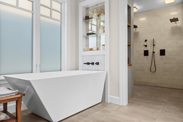 Modern bathroom featuring a white geometric bathtub, frosted windows, tiled floors, built-in shelving, and a walk-in shower with black fixtures and beige-tiled walls.