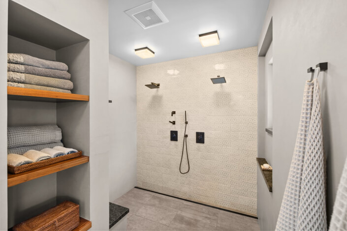 Modern bathroom featuring a double shower, beige textured wall tiles, two shower heads, a handheld shower, shelves neatly stocked with folded towels, and two towels hanging on a hook. The space is clean and minimalistic.