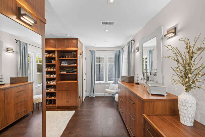 Spacious walk-in closet featuring wooden shelves and dressers, shoes arranged neatly, a large mirror, a white rug on the floor, and two windows with light blue curtains that allow natural light to fill the room.