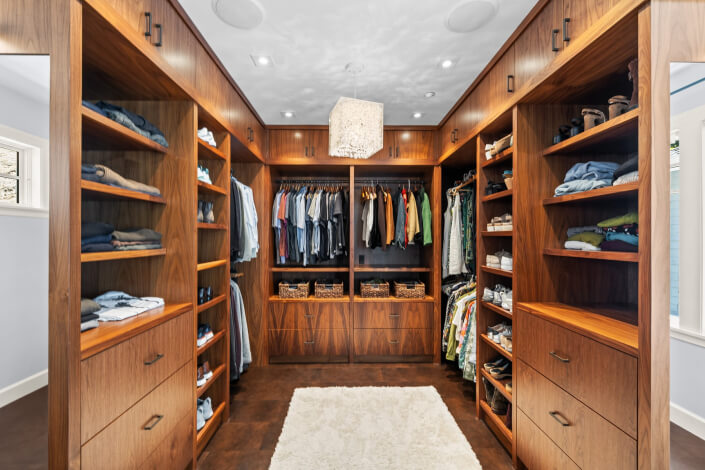 Spacious walk-in closet featuring wooden shelves and hanging rods, all neatly arranged with clothes and baskets. A white rug lies on the floor beneath a ceiling light fixture, highlighting the organized layout.