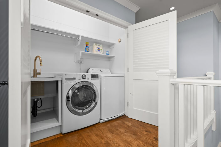 A modern laundry area featuring a front-load washer and a top-load dryer, complemented by white cabinets, shelves stocked with detergent, and a sink with a gold faucet. The walls are painted blue, and the space is finished with wood flooring and white trim.