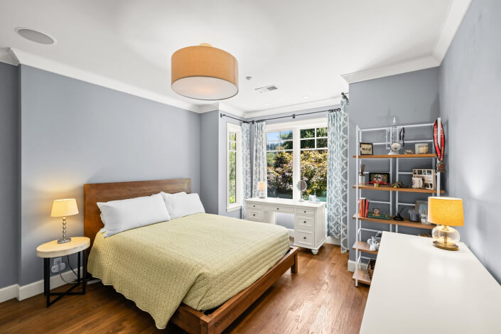 A bright bedroom with light blue walls, featuring a wooden bed covered in green bedding, two small lamps, a white desk situated beneath a window, and shelves displaying books and decor. The room has hardwood flooring and a visible ceiling light.