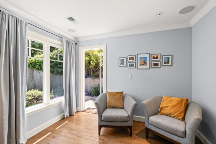A sunlit room with light blue walls features two gray armchairs topped with mustard yellow pillows, wood flooring, a large window dressed in gray curtains, a door that opens to the outdoors, and several pieces of framed art adorning the wall.