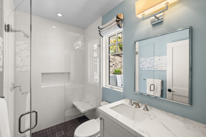 Modern bathroom featuring light blue walls, a glass-enclosed shower with a built-in shelf and small bench, a white vanity topped with marble, and a rectangular mirror. A window with a Roman shade lets in natural light, complemented by a wall-mounted light fixture.