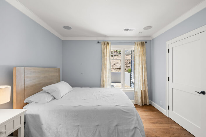 A minimal bedroom featuring a wooden floor, light blue walls, a bed with white bedding, a nightstand topped with a lamp, double doors draped with cream curtains, and a white closet door.