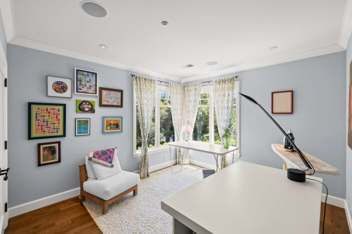 Bright home office and sewing room with blue walls, light wood floor, white desk, modern chair, large window with sheer curtains, wall art, and an ironing board in the corner. Sunlight fills the room, highlighting a vibrant and creative workspace.