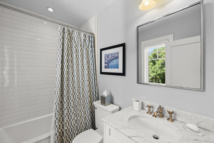 Bathroom featuring a white tile shower with a patterned curtain, marble countertop, rectangular mirror, framed art on the wall, tissue box placed on the toilet tank, and a window revealing green trees outside.