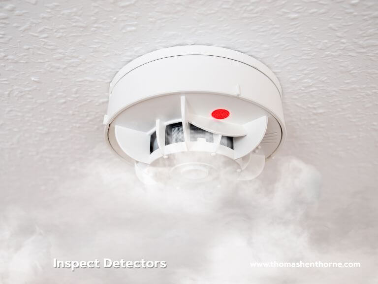 A smoke detector featuring a glowing red indicator is installed on a white ceiling, encircled by drifting smoke. The image contains the text “Inspect Detectors” along with a website URL.
