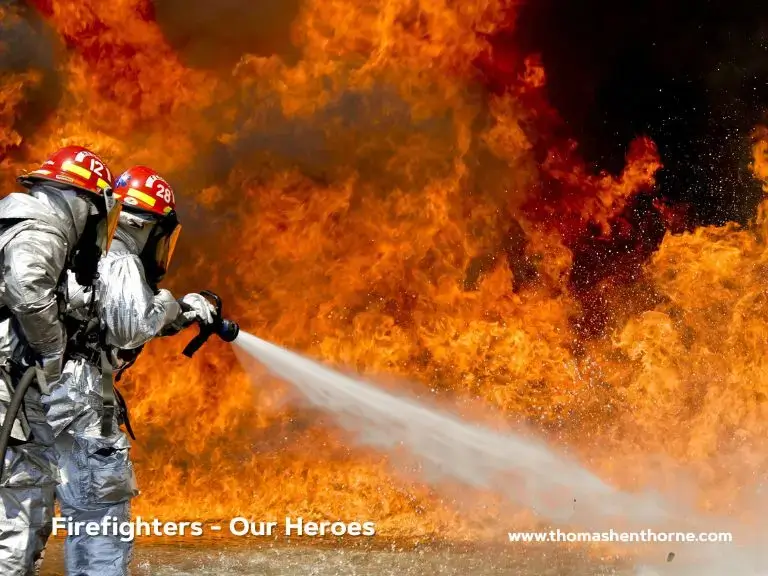 Two firefighters in silver protective suits aim water hoses at a massive fire, with bright flames soaring in the background. The image includes the phrase "Firefighters - Our Heroes" and features a website link in the lower right corner.