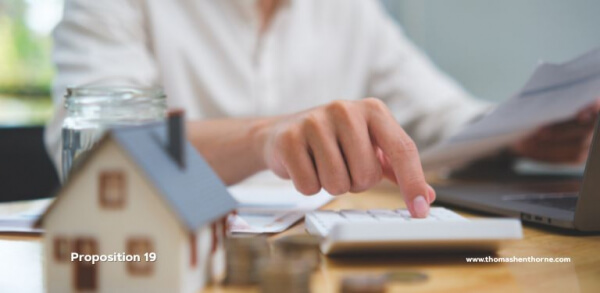 A person is seated at a desk using a calculator, surrounded by papers, coins, a glass of water, and a small model house, indicating they are engaged in financial or real estate calculations. The image includes the text "Proposition 19.