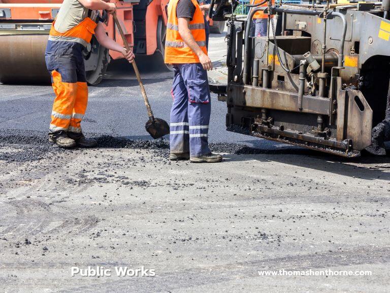 Two workers wearing safety vests and work pants use shovels to spread asphalt during road paving. Various pieces of road construction equipment, including a steamroller, appear in the background.