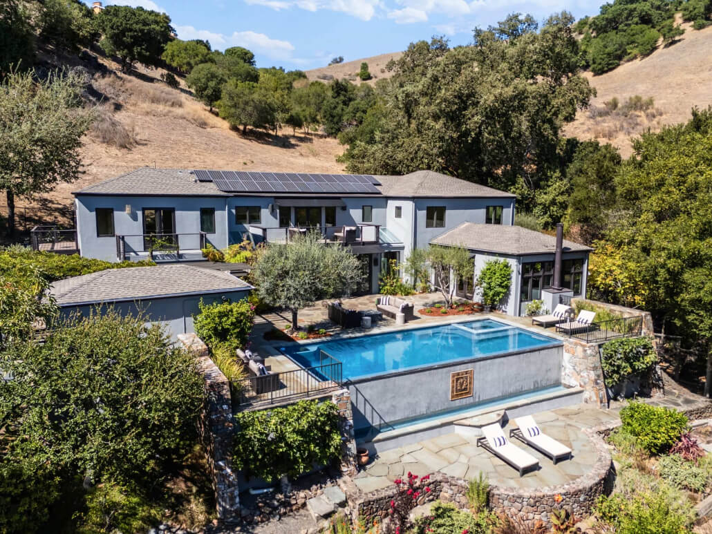 56 Miwok Dr_Large_003 HERO Modern two-story gray house featuring expansive windows, an infinity pool reflecting the sky, sleek lounge chairs, contemporary patio seating, and lush trees surrounding the hillside setting beneath a partly cloudy sky—an outstanding example of Marin real estate at 56 Miwok Drive, San Anselmo.