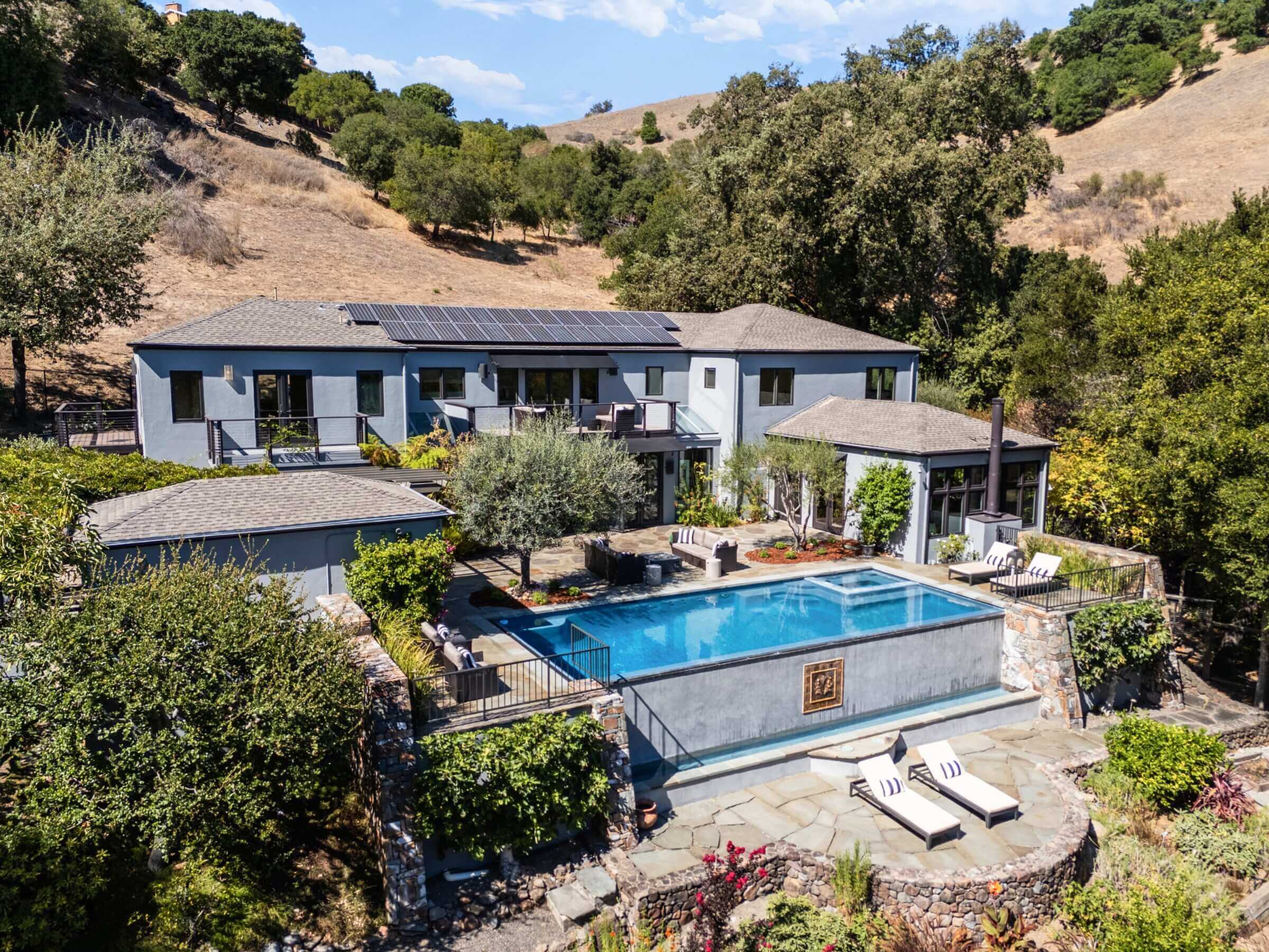 Modern two-story gray house featuring expansive windows, an infinity pool reflecting the sky, sleek lounge chairs, contemporary patio seating, and lush trees surrounding the hillside setting beneath a partly cloudy sky—an outstanding example of Marin real estate at 56 Miwok Drive, San Anselmo.